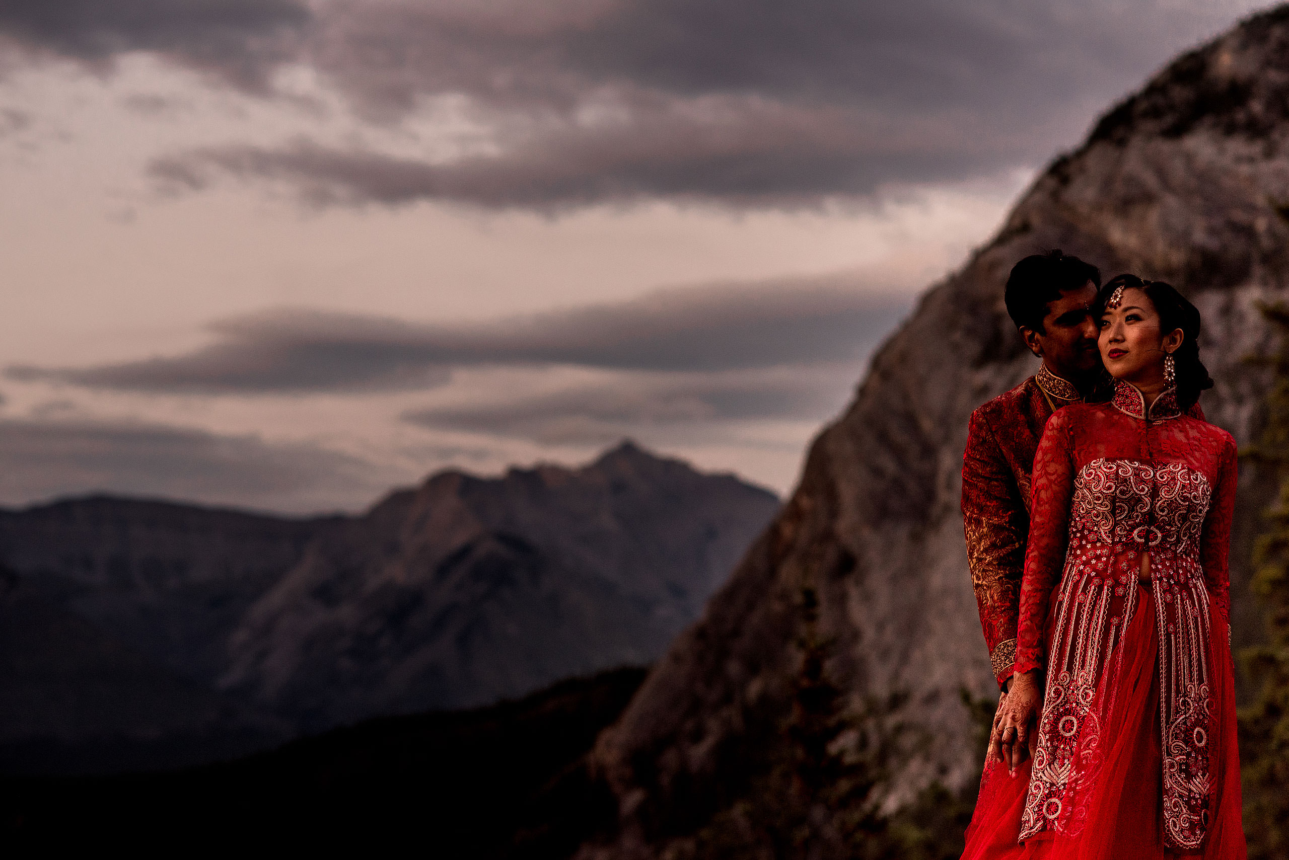 a bride and groom wearing red outfits with the mountains in the background an indian women in a red dress dancing for cascade ballroom banff springs wedding by sean leblanc
