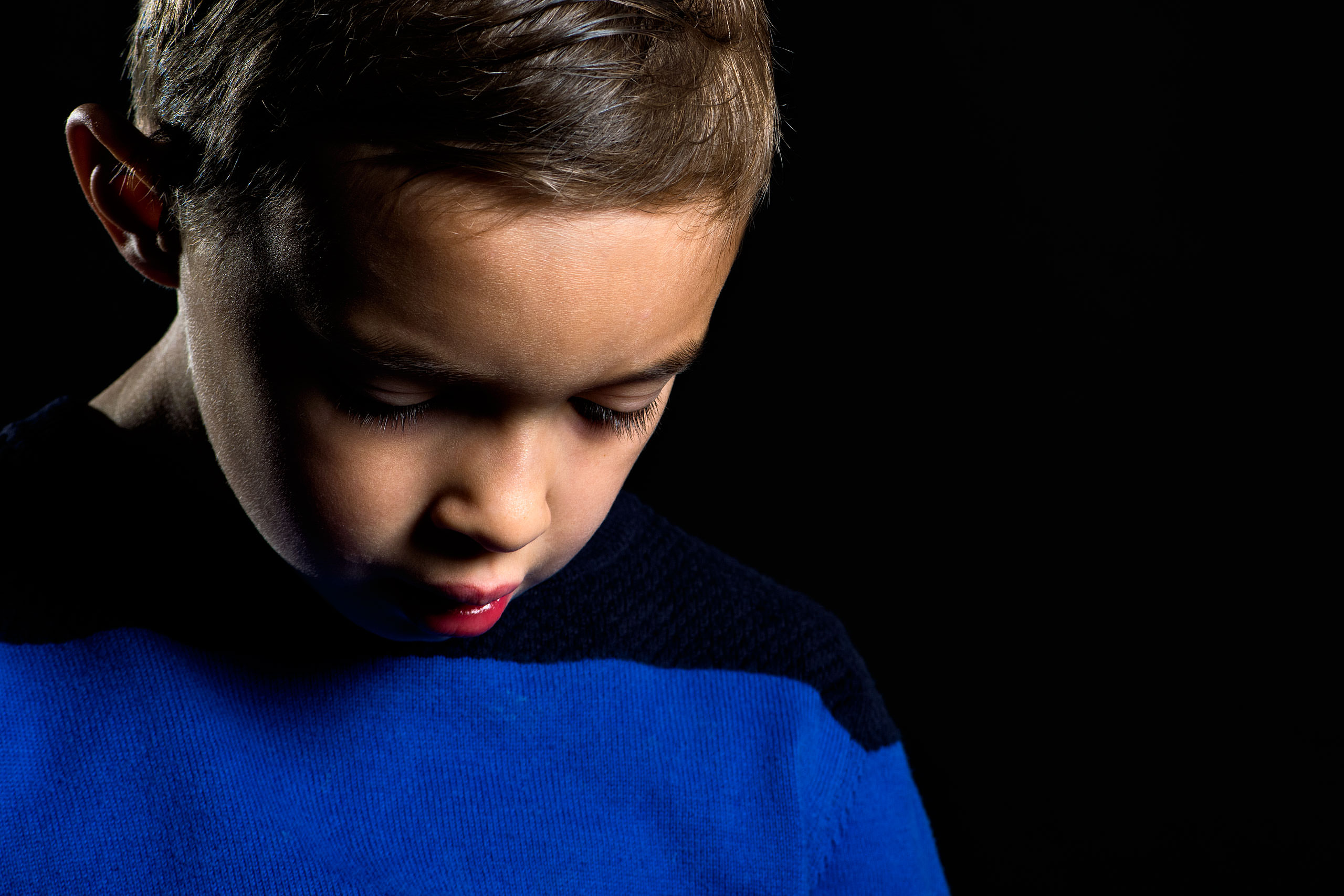 young boy looking towards the floor wearing a blue sweater on a black background by top family portrait photographer sean leblanc