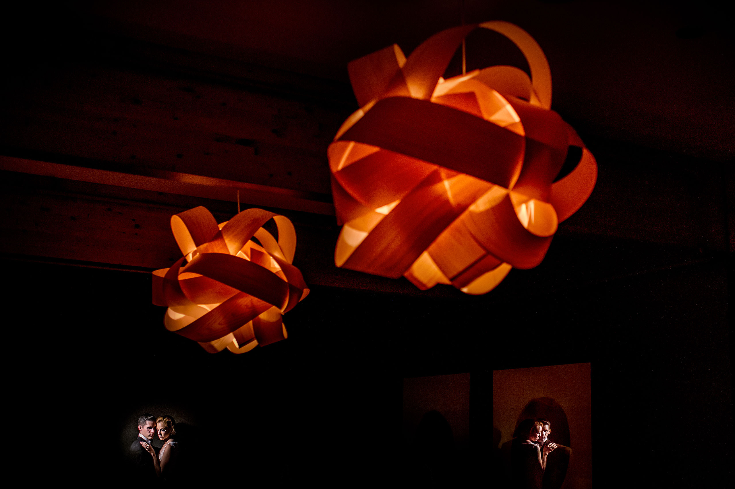 bride and groom embracing next to a wall with two large light fixtures in the foreground at Priddis Azuridge Wedding by sean leblanc