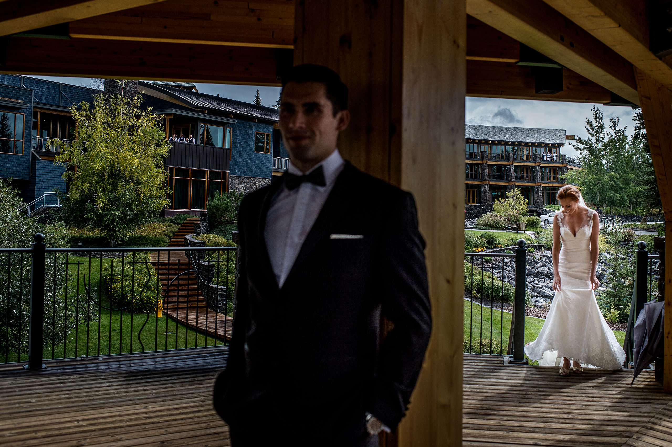 a bride walking up some stairs while her groom leans against a pole at Priddis Azuridge Wedding by sean leblanc