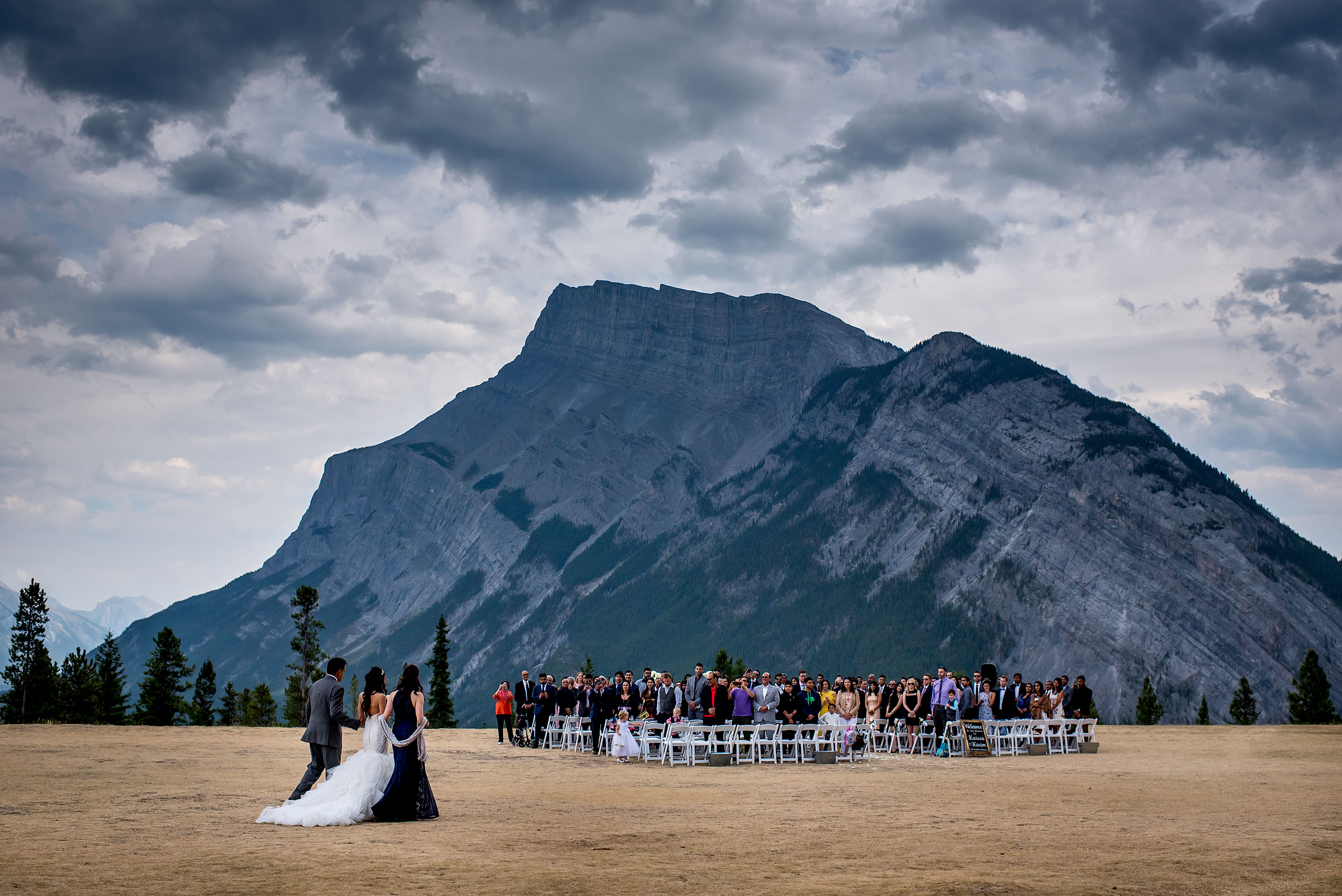 bride walking with parents to ceremony in front of Tunnel Mountain by a Banff wedding Photographer