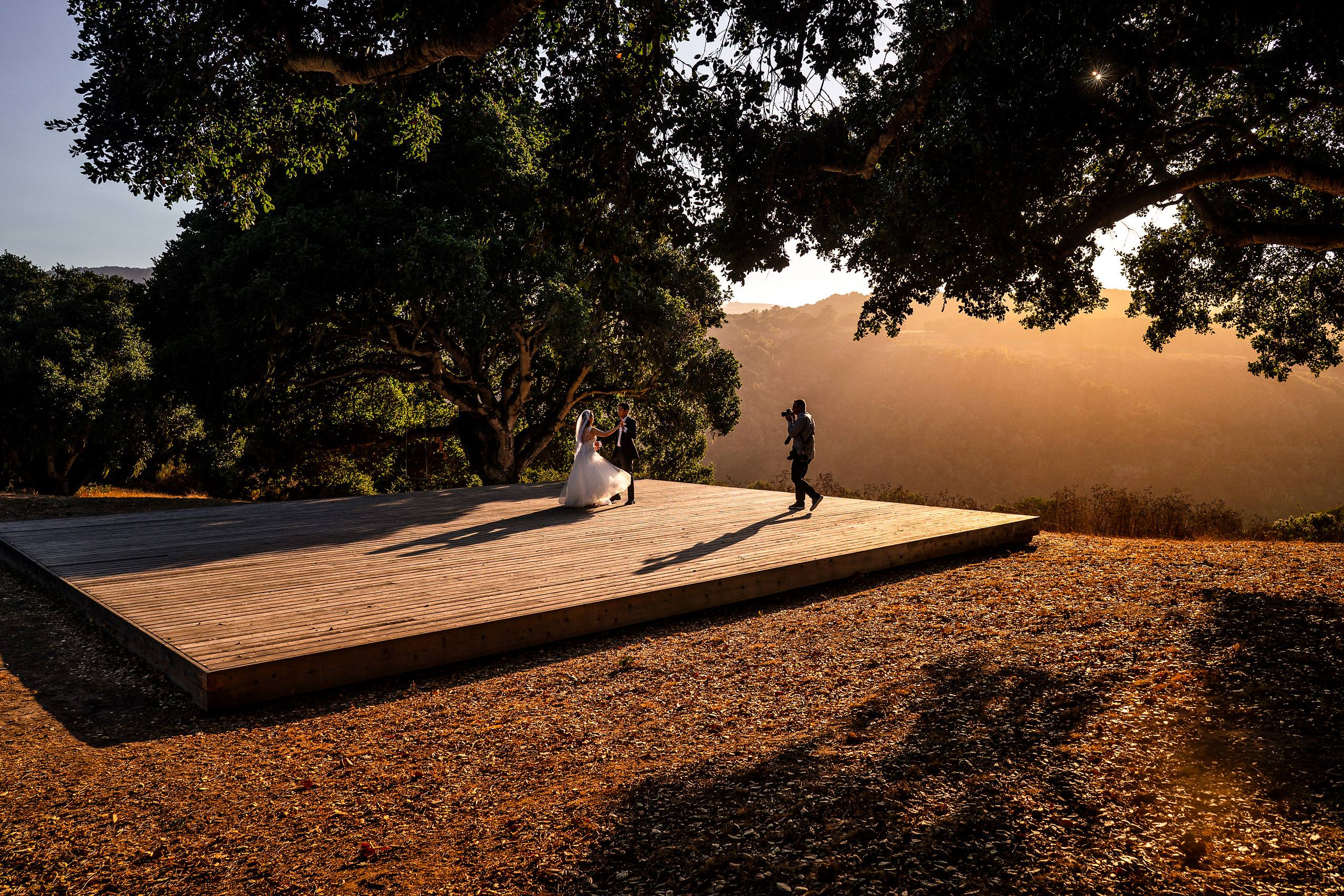 A photographer and a bride and groom on a wooden platform at sunset in California